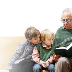 Older man sitting and reading a book to two young boys sat beside him on a wooden bench.